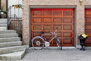A wooden garage door set in a brick house, with a bicycle parked in front and flower planters beside the entrance.