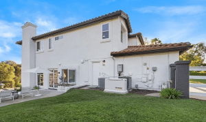 Mediterranean-style white two-story home with a tiled roof, green lawn, patio seating, and an outdoor Generac standby generator.