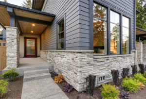 Modern home exterior with dark wood siding, large windows, stone veneer walls, a covered entryway, and landscaped garden beds.